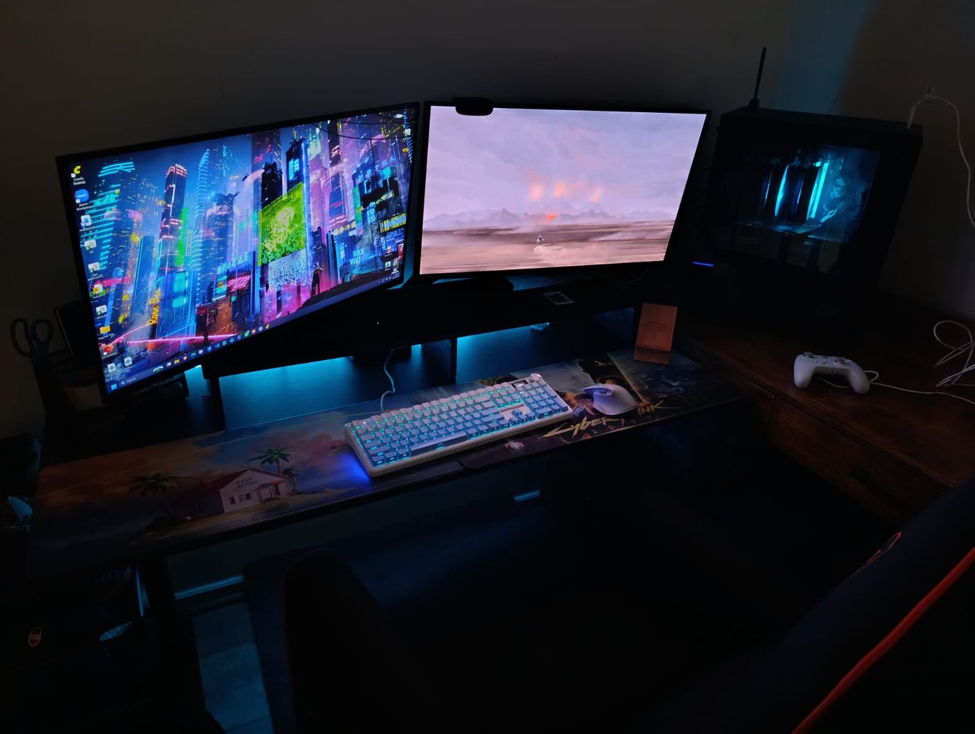 Jose's desk at night: two monitors, one with a neon cyberpunk city wallpaper, a white mechanical keyboard lit in cyan, a Cyberpunk 2077 desk mat and the PC glowing blue beside them